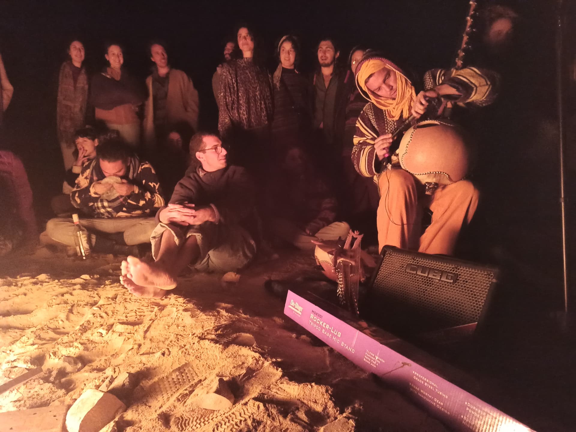 A night gathering in the desert around a musician playing a stringed gourd instrument